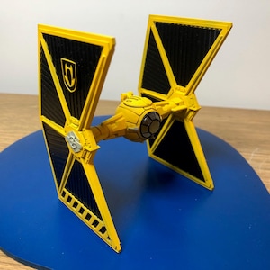 Puede incluir: Un modelo de caza TIE amarillo y negro, una nave espacial ficticia de una franquicia de ciencia ficción. El modelo tiene una sección de cabina central y dos alas de paneles solares. Las alas son negras con marcos amarillos y un emblema de escudo.
