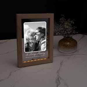 May include: A wooden framed photo with a black and white image of two people kissing. The frame has a warm light at the bottom and the text "In Loving Memory" is displayed. A vase with flowers is in the background.