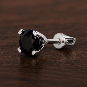 May include: A single stud earring featuring a round, faceted black gemstone held in a four-prong silver-toned metal setting. The earring includes a straight post and a flat, circular backing piece, showcasing a polished metallic finish throughout.