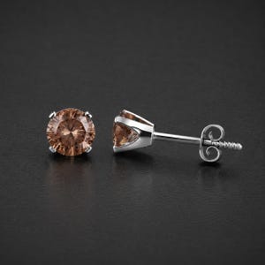 May include: A pair of stud earrings featuring round, light brown gemstones set in silver-colored metal. The earrings are displayed on a dark background, showcasing their sparkling appearance. The post and backing are also silver.