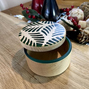 Puede incluir: Una pequeña caja redonda de madera con tapa. La tapa está decorada con un diseño de hojas verdes. El borde de la caja también está pintado de verde. La caja está sobre una superficie de madera.