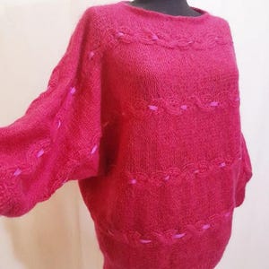 Può includere: Un maglione oversize color magenta con scollo tondo e maniche lunghe. Il maglione presenta fasce orizzontali decorative con un design a conchiglia e accenti di nastro rosa. La maglieria è esposta su un manichino.