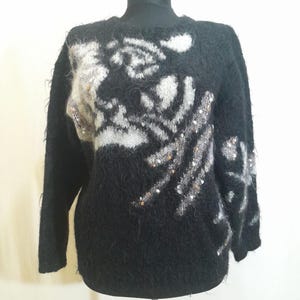 Op de afbeelding: Zwarte trui met een bloemenpatroon in wit en grijs, versierd met zilveren pailletten. De trui met lange mouwen wordt op een mannequin gepresenteerd. De trui heeft een fuzzy textuur.