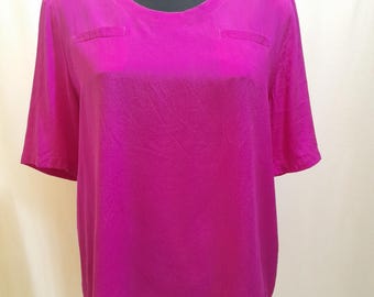 Op de afbeelding: Een fel magenta blouse met korte mouwen en een ronde halslijn. De top heeft twee kleine, rechthoekige zakken bij de schouders. De stof heeft een subtiele, getextureerde uitstraling en de blouse wordt getoond op een zwarte mannequin.