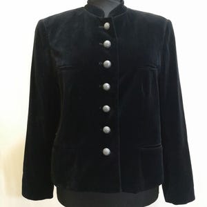 Op de afbeelding: Zwarte fluwelen jas met een opstaande kraag en zilverkleurige knopen. De jas heeft lange mouwen en twee voorzakken. Het kledingstuk wordt op een mannequin getoond.