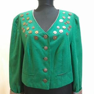 Puede incluir: Chaqueta verde con cuello en V y botones decorativos de color bronce. La chaqueta tiene una serie de adornos circulares de color bronce en los hombros y solapas. Los puños presentan una tela estampada. La chaqueta se exhibe en un maniquí negro.