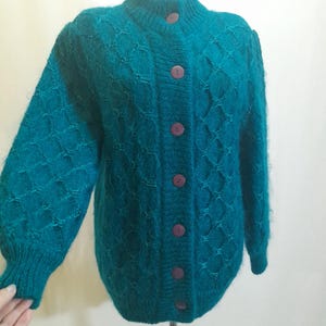 May include: Teal cardigan with a high collar and diamond pattern. The button-up sweater has long sleeves and is displayed on a mannequin. The buttons are a dark purple colour.