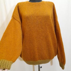 Puede incluir: Suéter de punto amarillo mostaza con cuello redondo y mangas largas. Los puños y el dobladillo son de color verde oliva. El suéter se exhibe en una cabeza de maniquí negra sobre un fondo blanco.