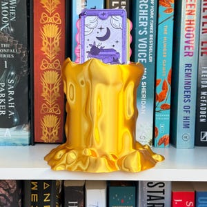 May include: A metallic gold, 3D-printed candle-shaped holder with a melted wax texture. Inside the holder sits a rectangular purple bookmark featuring a crescent moon, stars, and a stack of books illustration with the text "READ" at the bottom.