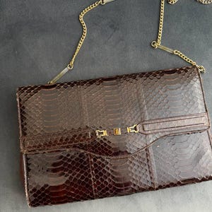 Può includere: Una pochette rettangolare marrone con un motivo a pelle di serpente. La borsa presenta una tracolla a catena color oro e una chiusura a fermaglio dorata. La pochette è in stile vintage, adatta per serate o occasioni speciali.