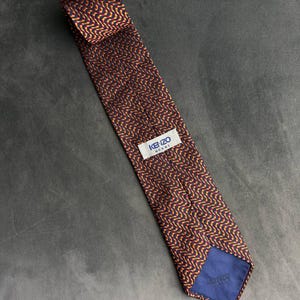 May include: A necktie featuring a repeating wavy pattern in shades of orange and yellow against a dark navy blue background. The tie includes a white fabric label stitched to the back with the text KENZO HOMME in blue, and a blue lining.