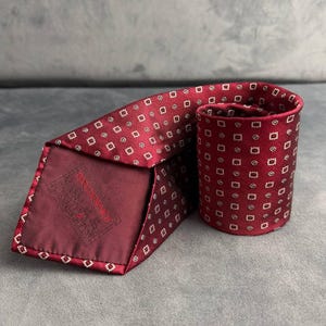 May include: A rolled burgundy tie with a repeating pattern of small white squares. The tie is made of a smooth fabric and has a label on the back. A classic accessory for formal wear.