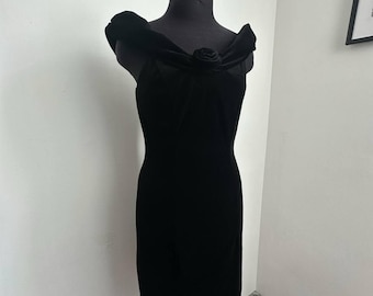 May include: A black velvet dress displayed on a mannequin. The dress features off-the-shoulder detailing and a rose embellishment at the neckline. The dress is knee-length and has a fitted silhouette, showcasing a classic, elegant design.