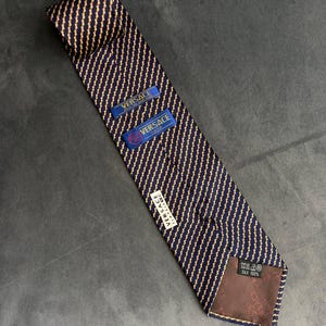 Pode incluir: Uma gravata azul-marinho com um padrão diagonal repetitivo de linhas douradas finas que lembram cordas. Inclui etiquetas azuis com o texto Versace Classic, uma etiqueta branca com Versace Classic e um forro castanho com Silk 100%.