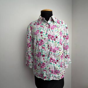 Peut inclure: Une chemise à manches longues boutonnée sur le devant, présentant un fond blanc avec un motif floral vif dans des tons magenta, rose et vert. La chemise possède un col classique, une patte de boutonnage et des poignets, présentée sur un mannequin.
