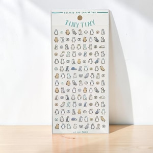 May include: A rectangular sticker sheet featuring numerous small, illustrated penguins in various poses and colors, including shades of gray, brown, and blue. The header text reads "RELAXED AND COMFORTING" and "TINY TINY," while the footer text reads "WE ARE PENGIN."