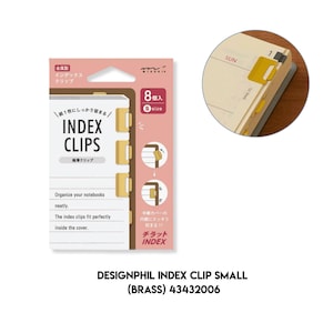 May include: A pack of eight small brass index clips by Midori, featuring a pink cardboard backing with instructions and text. The clips are shown attached to the edge of a notebook page in a circular inset, demonstrating their thin, flat design.