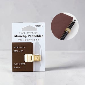 Op de afbeelding: Een bruine Midori Miniclip penhouder op een witte displaykaart met Japanse tekst. De penhouder heeft een goudkleurige metalen clip en een spiraalvormige veerlus voor een pen. Een cirkelvormige inzet toont de penhouder bevestigd aan een notitieboek.