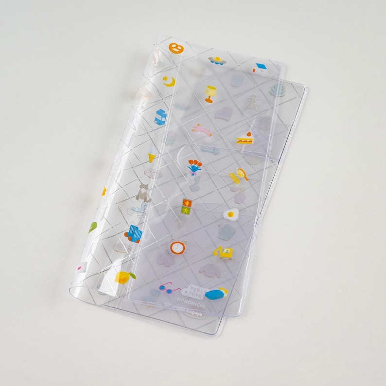 Puede incluir: Funda transparente de pl&aacute;stico para planificador con un patr&oacute;n de cuadr&iacute;cula e ilustraciones coloridas. La funda es rectangular y tiene un bolsillo. Las ilustraciones incluyen una luna, un pastel y una flor.