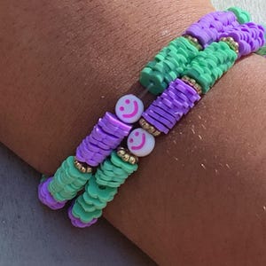 May include: Two bracelets made of green and purple beads with smiley face charms. The bracelets are stacked on a person's wrist.