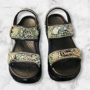 Op de afbeelding: Zwarte sandalen versierd met decoratieve elementen. De sandalen hebben een kroon, bloemmotieven en het woord "Smile" op één sandaal en "Chosen" op de andere. De bandjes zijn versierd met strass en parels.