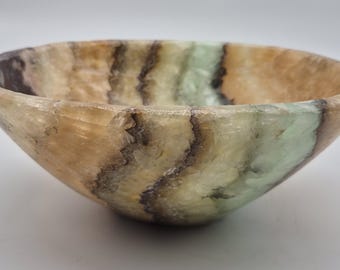 May include: A polished stone bowl featuring a natural banded pattern with translucent layers of amber, light green, and dark brown. The bowl has a smooth, rounded rim and a deep, curved basin, showcasing the organic mineral variations in the stone.