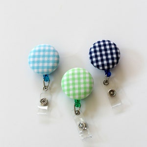 May include: Three fabric covered retractable badge holders with a checkered pattern. The holders are blue, green, and white.