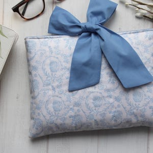 May include: A white and blue patterned fabric pouch with a blue bow. The pouch is made of a soft, quilted fabric and has a zipper closure.
