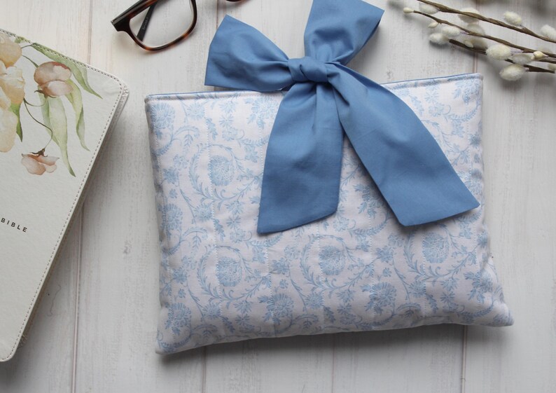 May include: A white and blue patterned fabric pouch with a blue bow. The pouch is made of a soft, quilted fabric and has a zipper closure.