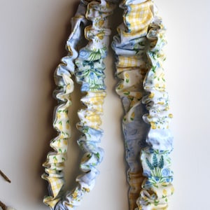 Puede incluir: Un cordón de tela con un diseño fruncido y con volantes, con un estampado de múltiples patrones de cuadros vichy amarillos, motivos florales azules y pequeñas flores amarillas. La cinta está unida a un cierre de metal plateado con una anilla.