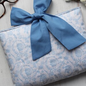 May include: A blue fabric pouch with a white floral pattern and a blue bow on top. The pouch is lying on a white wooden surface.