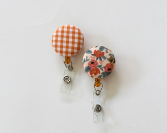 May include: Two fabric covered badge reels with clear plastic clips. One reel is orange and white gingham and the other is a floral pattern with pink, red, and green flowers on a cream background.