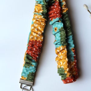 Puede incluir: Un cordón de tela con un diseño fruncido tipo coletero, con un patrón de patchwork de estampados florales y de cuadros en tonos amarillo mostaza, verde azulado, verde oliva y naranja óxido, acabado con un cierre y anilla de metal plateado.