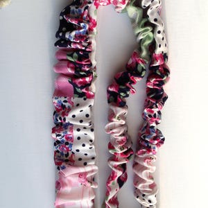 Puede incluir: Un cordón de tela con un diseño fruncido y elástico, con un patrón de retazos de flores y lunares en tonos rosa, blanco, azul marino y verde. Un cierre de metal plateado y una anilla circular están sujetos en el extremo inferior.