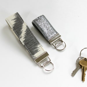 May include: Two keychains with silver key rings and a set of three keys. One keychain has a gray and white patterned fabric strap and the other has a silver glitter fabric strap.