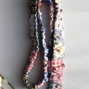 Puede incluir: Un cordón de tela con un diseño de patchwork que presenta varios patrones florales, de cuadros vichy y lisos en tonos rosa, azul, verde y blanco. La correa está fruncida con elástico y unida a un cierre de metal plateado con una anilla redonda.