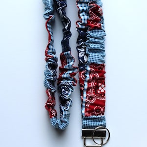 Puede incluir: Un cordón de tela con un diseño fruncido y elástico que presenta un patrón de retazos en colores rojo, blanco y azul. La tela muestra secciones alternas de estampado de cachemira rojo, cuadros vichy azul claro y flores azul oscuro.