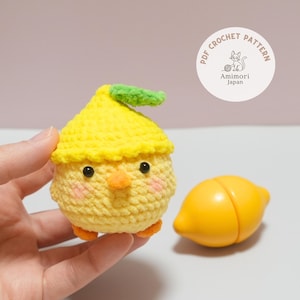 Könnte beinhalten: Ein handgehäkeltes gelbes Küken mit einem grünen Blatt auf dem Kopf, schwarzen Augen und einem orangefarbenen Schnabel. Eine gelbe Spielzeugzitrone befindet sich neben dem Küken. Das Bild enthält einen weißen Kreis mit dem Text "PDF Crochet Pattern" und "Amimori Japan".