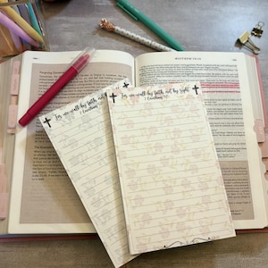 May include: An open Bible with pink tabs, a red pen, and two floral-patterned notepads with the text "For we walk by faith, not by sight." Pens and a paper clip are also visible. The notepads have lined pages.
