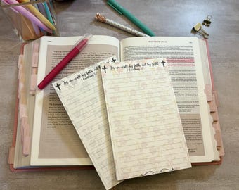 May include: An open Bible with pink tabs, a red pen, and two floral-patterned notepads with the text "For we walk by faith, not by sight." Pens and a paper clip are also visible. The notepads have lined pages.
