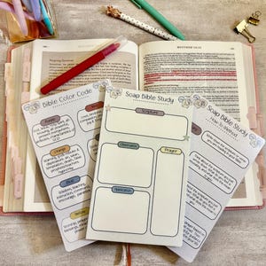 May include: An open Bible with a red pen, surrounded by study guides. The guides include "Bible Color Code" and "Soap Bible Study" with sections for scripture, observation, and prayer. Pens and a paper clip are also visible.