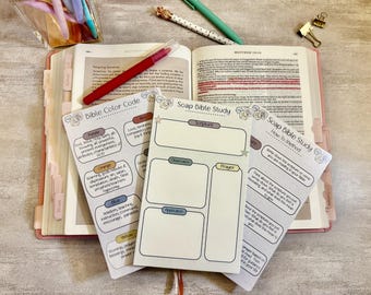 May include: An open Bible with a red pen, surrounded by study guides. The guides include "Bible Color Code" and "Soap Bible Study" with sections for scripture, observation, and prayer. Pens and a paper clip are also visible.