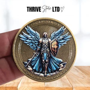 May include: An antique gold-coloured coin depicting the Archangel Michael. The coin shows Michael with blue wings, a shield, and a sword. The text "THE ARCHANGEL" and "MICHAEL WATCHES OVER US" are inscribed around the edges.