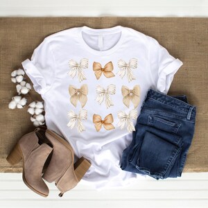 May include: A white t-shirt featuring a pattern of brown and beige watercolour bows. The shirt is displayed on a brown hessian surface with a pair of brown ankle boots and blue jeans.