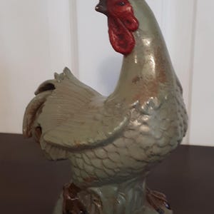 May include: A green ceramic rooster figurine with a red comb and wattle. The rooster is standing on a base with leaves and has a textured surface.