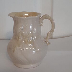 May include: A small, cream-colored ceramic creamer with a delicate floral design. The creamer has a rounded body and a curved handle.