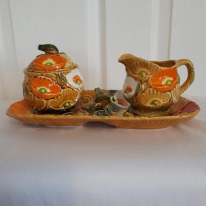 May include: A ceramic sugar and creamer set with a tray. The set features a floral design with orange and white poppies on a yellow background.