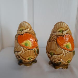 May include: A pair of ceramic salt and pepper shakers. Each shaker is egg-shaped, with a tan base and a vibrant orange poppy flower design. The flowers have yellow centers and green accents. The shakers sit on a dark green base.