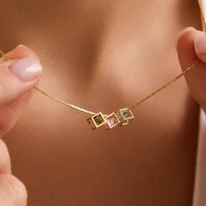 Puede incluir: Un delicado collar de oro con tres pequeños colgantes cuadrados con piedras preciosas de colores. El collar se sostiene, mostrando el diseño y el sutil brillo de las gemas. La cadena es fina y elegante, perfecta para el uso diario.