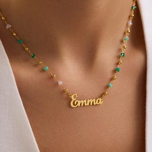 May include: A gold-colored necklace with the name "Emma" in a cursive font. The necklace features small, colorful beads in green, pink, and white. The necklace is worn around the neck.